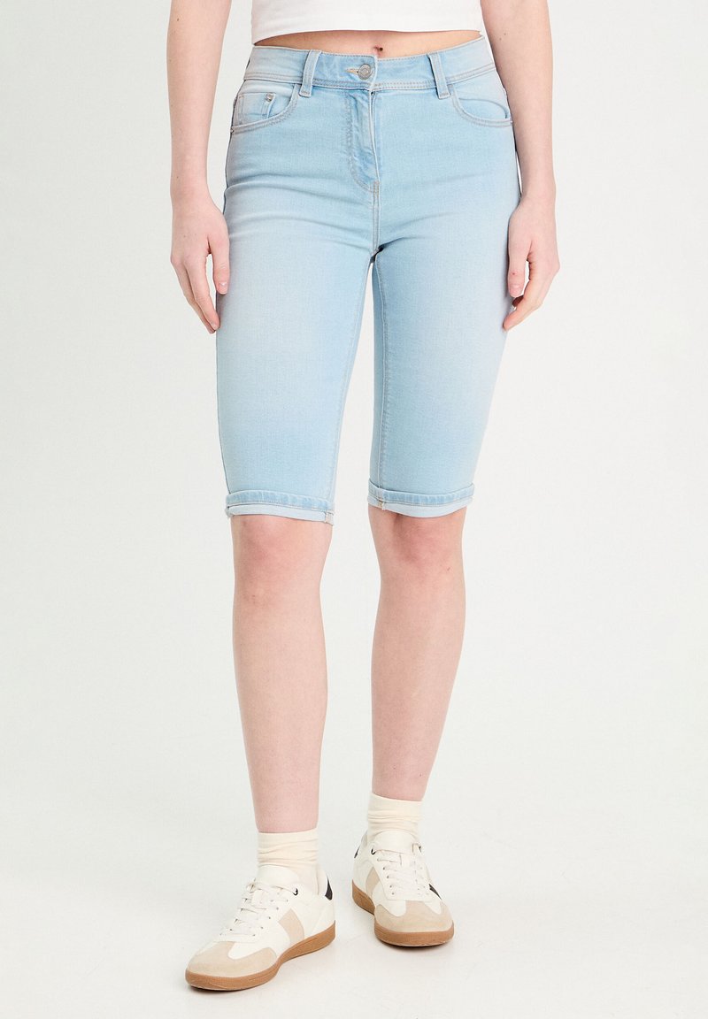 Bermuda shorts en denim bleu clair avec une coupe ajustée, ourlets roulés et deux poches avant, assortis à des baskets blanches et des chaussettes beiges.