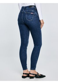 Naine, kes kannab tumedat sinist kitsaid teksaseid ja musta madala kontsaga rihmikkingi, seisab ühevärvilise heleda tausta ees.