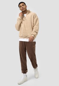 Beige fleecetrøye med halv glidelås og logo-detalj, kombinert med brune fløyelsjoggebukser og hvite sneakers.