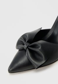 Schwarzer Leder-Schuh mit spitzer Kappe, verziert mit einer großen Schleife vorne, mit glatter Textur und schlanker Silhouette.