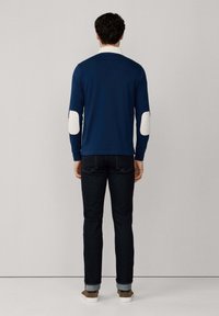 Mann steht mit dem Rücken zu sehen und trägt ein marineblaues Langarmhemd mit weißen Ellbogenpatches, dunkle Jeans und braune Sneaker mit weißen Sohlen.