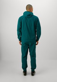 Set di giacca e pantaloni impermeabili color teal. La giacca ha un cappuccio, polsini elastici e una vestibilità comoda; i pantaloni presentano orli elastici e un design a gamba dritta.
