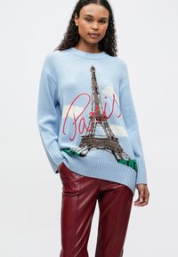 Frau in hellblauem Pullover mit dem Eiffelturm und rotem "Paris"-Text, kombiniert mit dunkelroten Lederhosen, stehend vor einem weißen Hintergrund.
