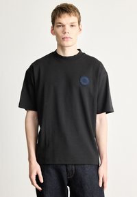 Μαύρο βαμβακερό μπλουζάκι με στρογγυλή λαιμόκοψη, κοντά μανίκια και κυκλικό μπλε σχέδιο στον αριστερό θώρακα.