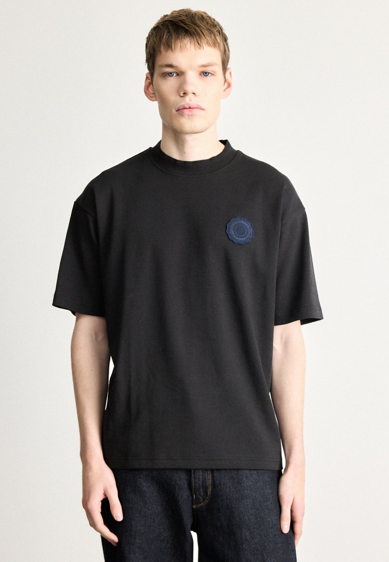 Μαύρο βαμβακερό μπλουζάκι με στρογγυλή λαιμόκοψη, κοντά μανίκια και κυκλικό μπλε σχέδιο στον αριστερό θώρακα.