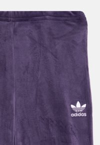 Violet velūro kelnės su lygiu paviršiumi, kuriose apačioje kairėje pusėje yra nedidelis baltas adidas logotipas.