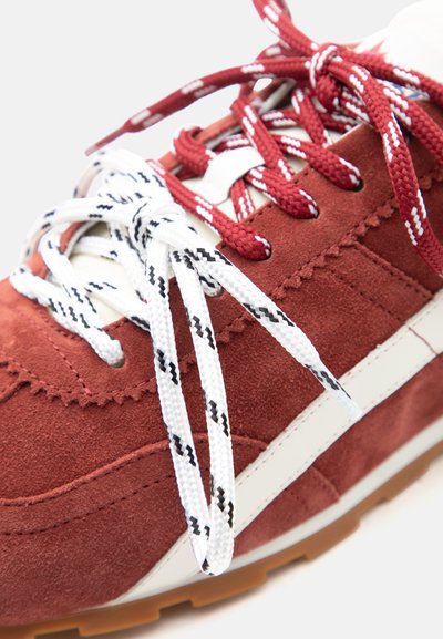 Gros plan sur une basket en daim rouge avec une bande latérale blanche, des lacets blancs avec des détails noirs, et des lacets rouges noués sur le dessus.