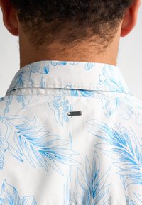 Camicia a maniche corte con tessuto bianco decorato da motivi floreali blu, colletto classico e una piccola etichetta metallica sullo scollo.
