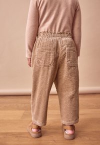 Beige fløjlsbukser med en afslappet pasform, der har to baglommer og en tekstureret overflade, kombineret med brune sandaler på et trægulv.