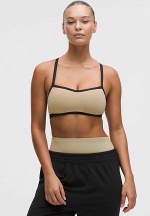 Frau trägt einen beigen und schwarzen Sport-BH und schwarze Hose mit beigem Bund, steht vor einem schlichten hellen Hintergrund.
