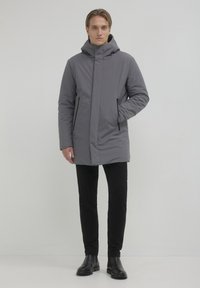 Cappotto grigio imbottito con colletto alto e cappuccio, dotato di due tasche laterali con zip e una linea dritta. Indossato sopra pantaloni neri slim-fit.