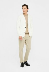 Bela pletena jopica z rjavimi gumbi, bež zgornji del, bež hlače in črni čevlji. Model stoji na nevtralni podlagi.