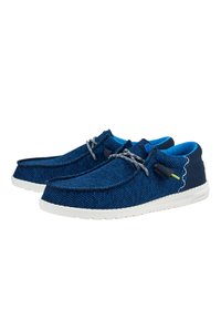 Mocasines de tela azul con una superficie texturizada, cordones elásticos, suelas contrastantes en blanco y un forro interno de azul claro. Detalle de costura decorativa.