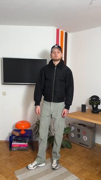 Černá bomber bunda se zipem, velkými předními kapsami a vysokým límcem. Nošena s světle šedými kalhotami a teniskami, v místnosti s nábytkem a dekoracemi.