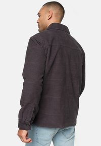 Dunkelgrå fløjlsjakke med krave, knaplukning ved ærmerne og lodrette ribbede strukturer, set fra ryggen, parret med lyseblå jeans.