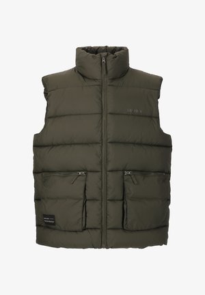 Gilet matelassé vert olive avec un col haut, deux poches avant, porté sur une chemise à manches longues noire et associé à un pantalon noir et des baskets.
