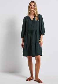 Kerudung bertingkat berwarna hijau tua terbuat dari kain ringan dengan leher V dan lengan tiga perempat. Memiliki plakat kancing dan potongan yang longgar.