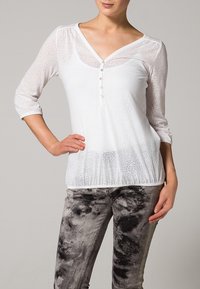 Weiße, halbtransparente Bluse mit Dreiviertelärmeln, einem V-Ausschnitt und vier Knopfakzenten. Kombiniert mit grauen Jeans im Batikmuster.
