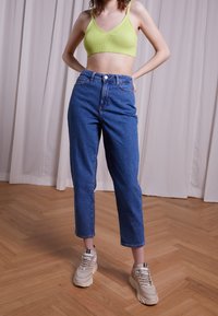 Vyšší modré džíny s volným střihem, spárované se žebrovaným limetkově zeleným crop topem a béžovými teniskami. Pozadí tvoří přírodní dřevěná podlaha.