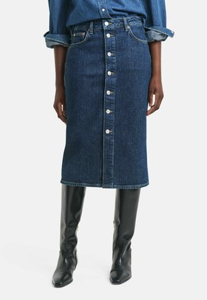 Jupe crayon en denim foncé avec une fermeture à boutons sur le devant, coupe mi-longue et légère décoloration. Associée à des bottes en cuir noires jusqu'aux genoux.