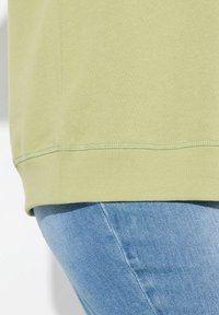 Felpa verde chiaro con orlo a coste, posizionata sopra un denim blu. Il tessuto appare morbido con una texture liscia.