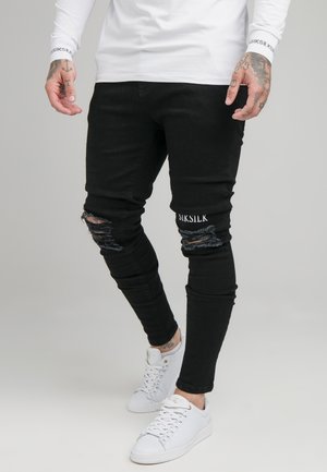 Mężczyzna noszący czarne, podziurawione obcisłe jeansy z logo SikSilk oraz białe trampki, pokazany od talii do stóp na szarym tle.