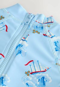 Ljusblå zip-up baddräkt med nautisk design som har båtar och moln. Tillverkad av elastisk tyg med en mjuk textur.