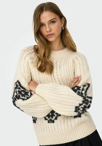 Dikke gebreide trui in crème met zwarte geometrische patronen op de mouwen, voorzien van een ronde halslijn en een geribbelde textuur.