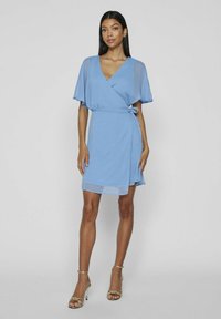 Svetlomodré zavinovacie šaty s krátkymi volánikmi, jemnou textúrou, dĺžkou po kolená a viazačkou v páse. Zlaté sandále na podpätku.