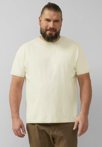 Kremowa koszulka z krótkim rękawem, wykonana z bawełny, z okrągłym dekoltem i luźnym krojem. Nosi ją z brązowymi spodniami, prezentując ją na neutralnym tle.