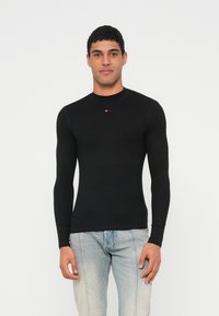 Schwarzes, langärmeliges, körpernah geschnittenes Shirt mit geripptem Muster, rundem Ausschnitt und kleinem Logodetail auf der Brust, kombiniert mit hellblauen Jeans.