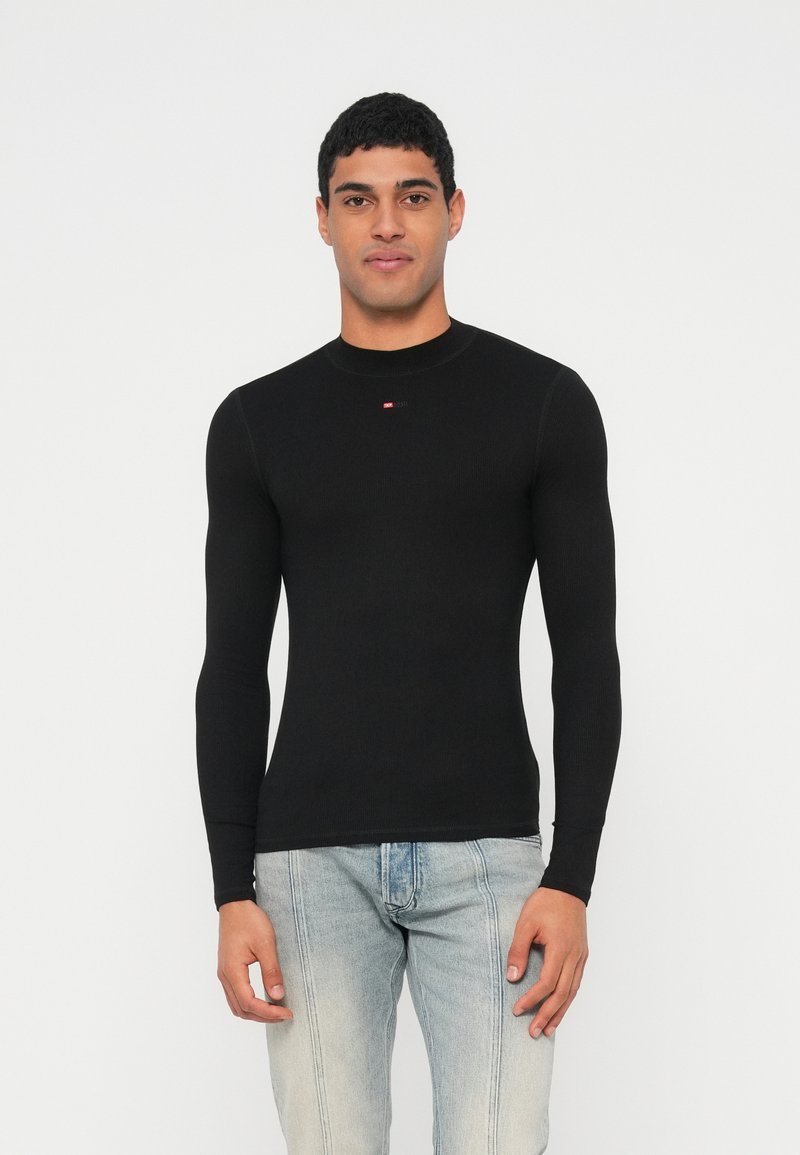 Schwarzes, langärmeliges, körpernah geschnittenes Shirt mit geripptem Muster, rundem Ausschnitt und kleinem Logodetail auf der Brust, kombiniert mit hellblauen Jeans.