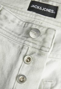 Bela denim tkanina z blagim diagonalnim vzorcem, opremljena z črnim oznako JACK & JONES, srebrnimi gumbi in graviranimi podrobnostmi blagovne znamke.