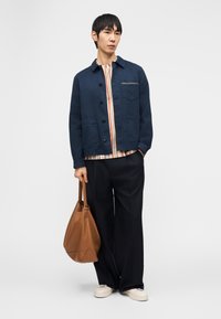 Férfi tengerészkék dzsekiben, csíkos ingben, fekete bőszárú nadrágban, fehér sportcipőben, kezében nagy barna bőrből készült válltáskával, egyszínű háttér előtt áll.