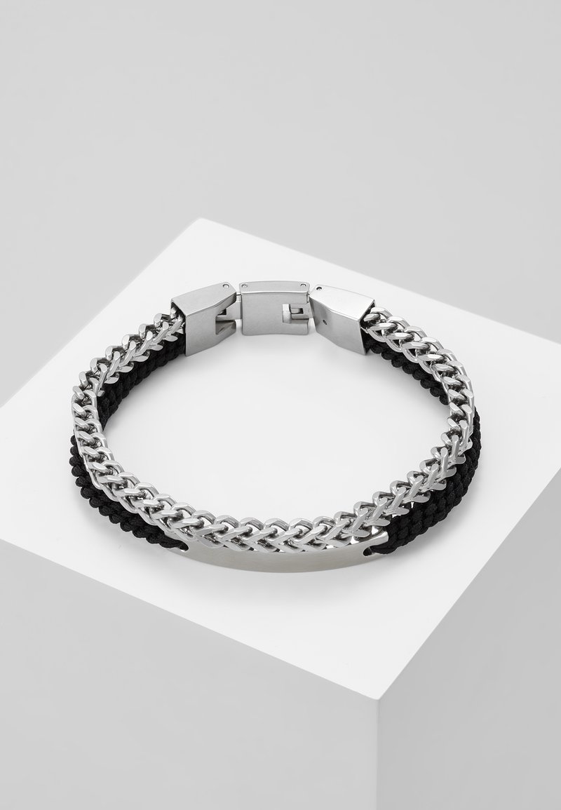 Bracelet en chaîne argentée et cordon noir tressé avec fermoir en métal poli et plaque rectangulaire vierge sur un socle blanc.