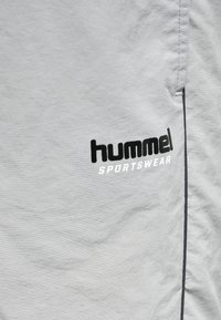 Lichtgrijze sportkledingstof met een gestructureerde afwerking, voorzien van een zwart "hummel" logo en "SPORTSWEAR" tekst in het wit, aan de linkerkant.