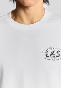 Witte katoenen t-shirt met een ronde halslijn en een geprinte grafiek in grijs en zwart met de tekst "L&S" binnen een ovale vorm.