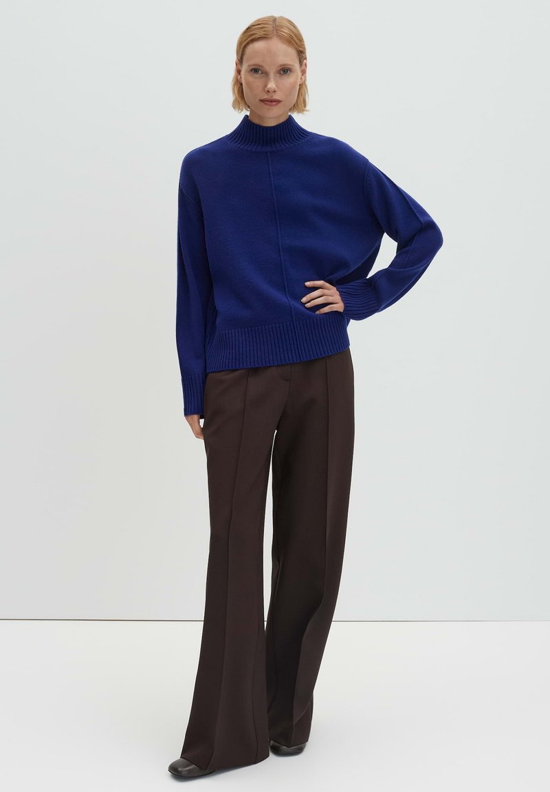 Pull à col roulé tricoté bleu roi avec larges manches, associé à un pantalon marron taille haute. Texture lisse et coupe décontractée.