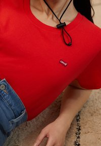 T-shirt rouge à manches courtes en coton, comportant un petit patch logo Levi's. Porté avec un jean en denim taille haute et un cordon noir.