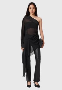 Mustavartaloinen toppi, jossa on läpinäkyvä kangas, epäsymmetrinen helma ja pitkä hiha, yhdistettynä istuviin mustiin housuihin ja tekstuuriin mustiin korkokenkiin.