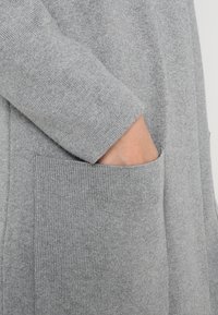 Grau gestrickte Strickjacke mit einem strukturierten Stoff, langen Ärmeln und übergroßen Fronttaschen. Glatte Oberfläche mit sichtbaren Nähten.