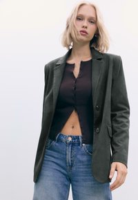 Mørkegrøn blazer lavet af blødt materiale, lagdelt over en brun ribbet top med knapper. Båret med lys blå højtaljede jeans.