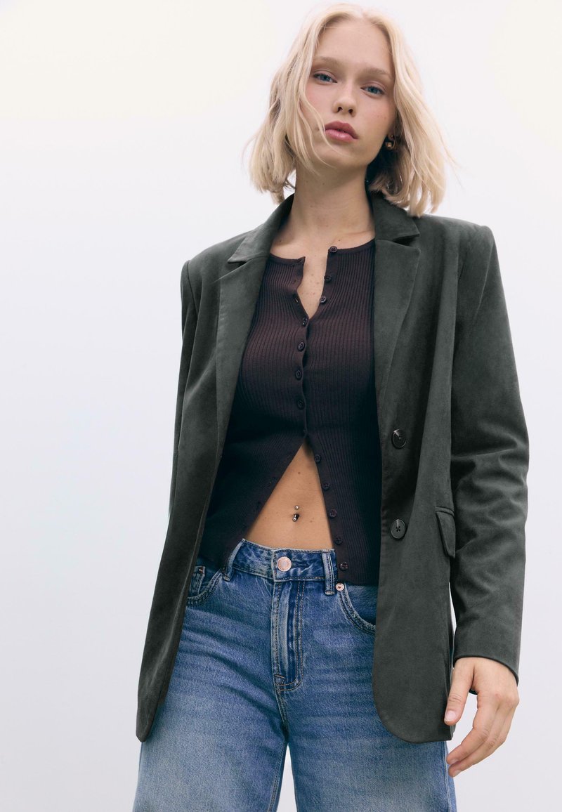 Mørkegrøn blazer lavet af blødt materiale, lagdelt over en brun ribbet top med knapper. Båret med lys blå højtaljede jeans.
