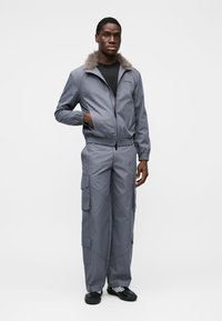 Γκρι ζακέτα με φερμουάρ και ψεύτικη γούνα στο γιακά, σε συνδυασμό με γκρι παντελόνι cargo με πολλές τσέπες. Το μοντέλο φοράει μαύρο πουκάμισο και παπούτσια.