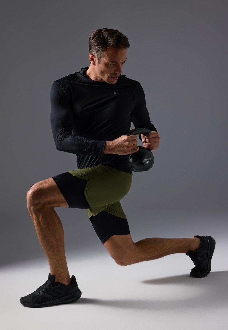 Czarna, długorękawowa koszulka sportowa, oliwkowozielone i czarne spodenki, trzymający czarny kettlebell, w pozycji wykroku na szarym tle.