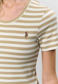 Svītrots ribots adīts tops bēšā un krēmkrāsā ar apaļu izgriezumu, ar izšuvumu augšējā kreisajā stūrī. Īsas piedurknes.