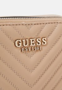 Beige quiltad läderplånbok med en guldfärgad "GUESS"-logotyp och inskriptionen "EST 1981", med synliga sömmar och dragkedjestängning.