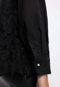 Blusa preta com detalhes em renda floral, fecho de botão na manga, textura de tecido suave e uma silhueta estruturada.