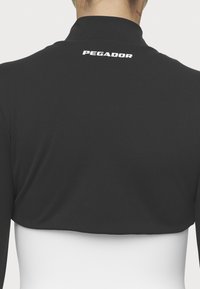 Melnā sporta jakas ar īsu piegriezumu un augstu kaklu. Aizmugurē ir balts uzraksts "PEGADOR". Gluda tekstūra un pieguļošs dizains.