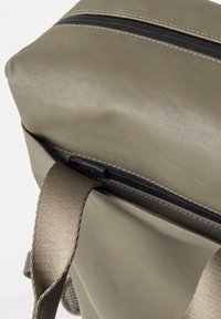 Zaino verde oliva con superficie testurizzata, dotato di una cerniera nera e spallacci beige. Design compatto con parte superiore arrotolata e tasca laterale.